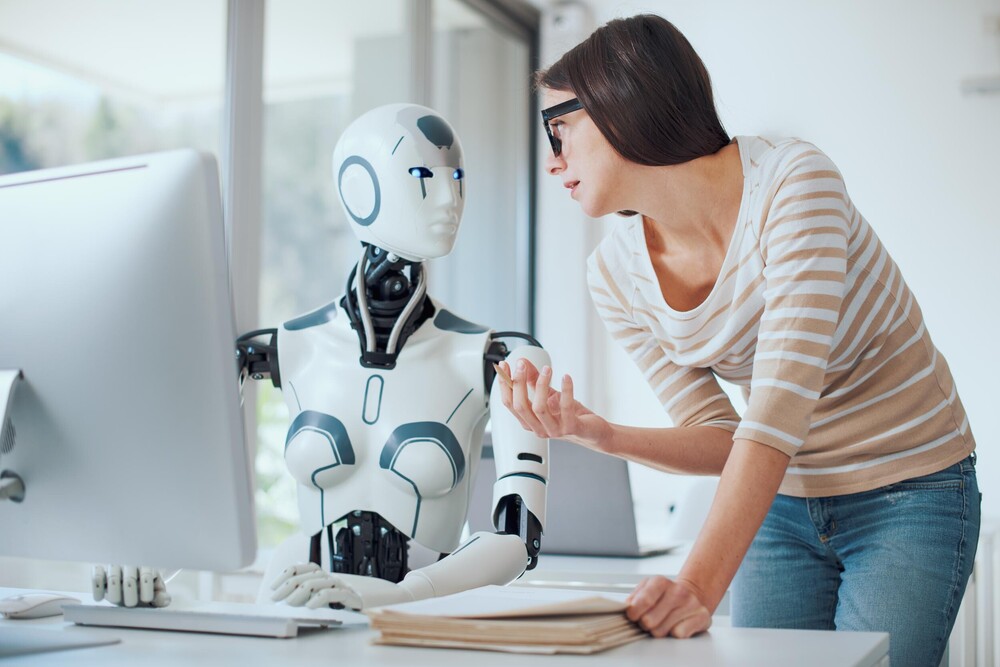Eine Frau erklärt etwas einem an einem Schreibtisch sitzenden Roboter